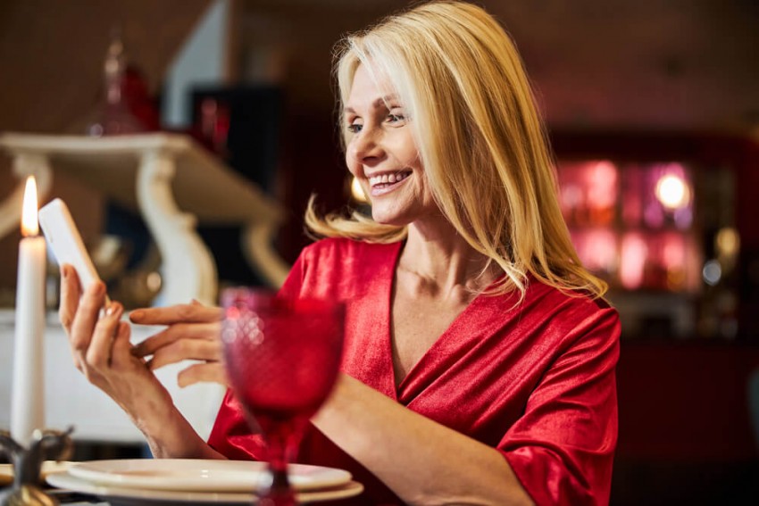 En smilende moden blond kvinde kigger på sin smartphone under en romantisk middag.